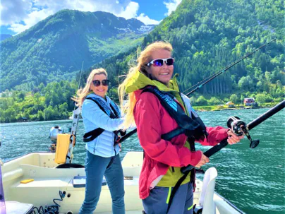 Two smiling women in life vests are fishing from a boat on a sunny day in a Norwegian fjord with steep green mountains in the background.