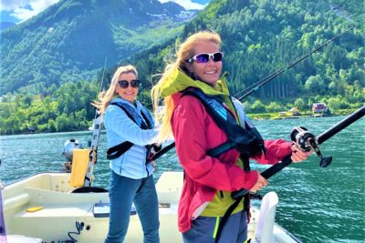 Two smiling women in life vests are fishing from a boat on a sunny day in a Norwegian fjord with steep green mountains in the background.